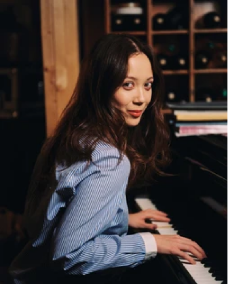 Laufey, the 2026 honorary chair of Library Card Sign-up Month, sits at a piano and looks over her shoulder toward the camera.